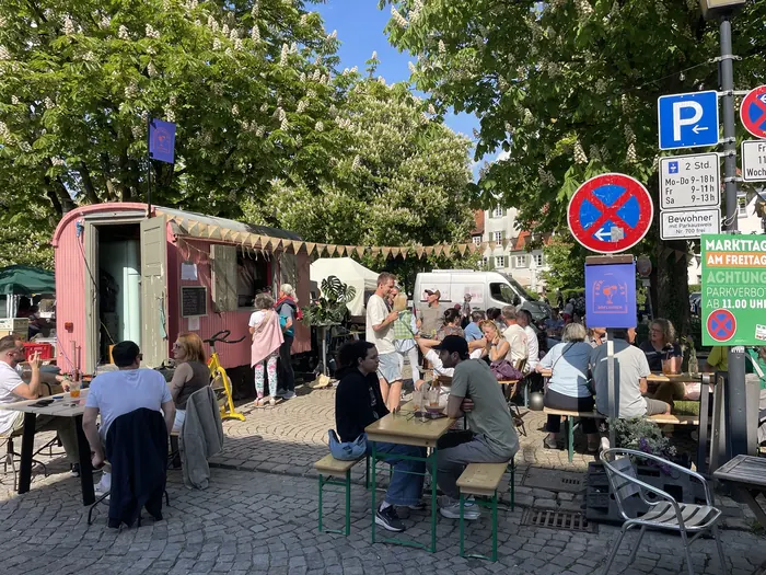 Gäste an Biertischen vor dem Bauwagen unter blühenden Kastanien
